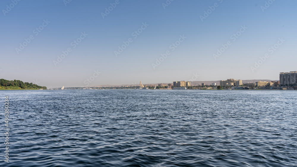 The wide blue Nile flows calmly. Ripples on the water. Felucca can be seen in the distance. On the banks - city houses, green vegetation. Clear azure sky. Egypt
