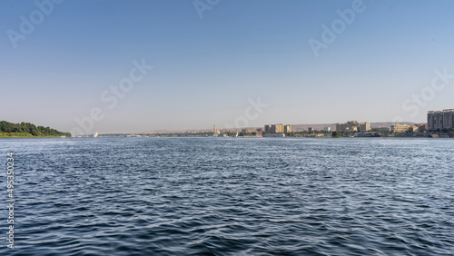 The wide blue Nile flows calmly. Ripples on the water. Felucca can be seen in the distance. On the banks - city houses, green vegetation. Clear azure sky. Egypt