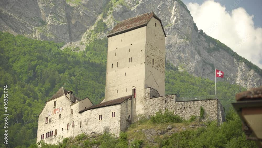 Low angle of historical Sargans Castle with Swiss flag waving on a hill near Mount Gonzen surrounded by dense green pine forest, Switzerland Alps Low angle of historical Sargans Castle with Swiss flag waving on a hill near Mount Gonzen surrounded by dense green pine forest, Switzerland Alps