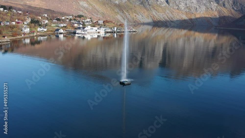 A fountain in the fjord near the fishing village of Torsken, Norway. The sky is reflected in the mirrorlike still waters. Mountains tower in the background. Slow-motion, orbit, aerial. photo