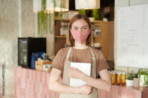 Horizontal medium portrait of young waitperson wearing apron and protective mask holding digital tablet looking at camera