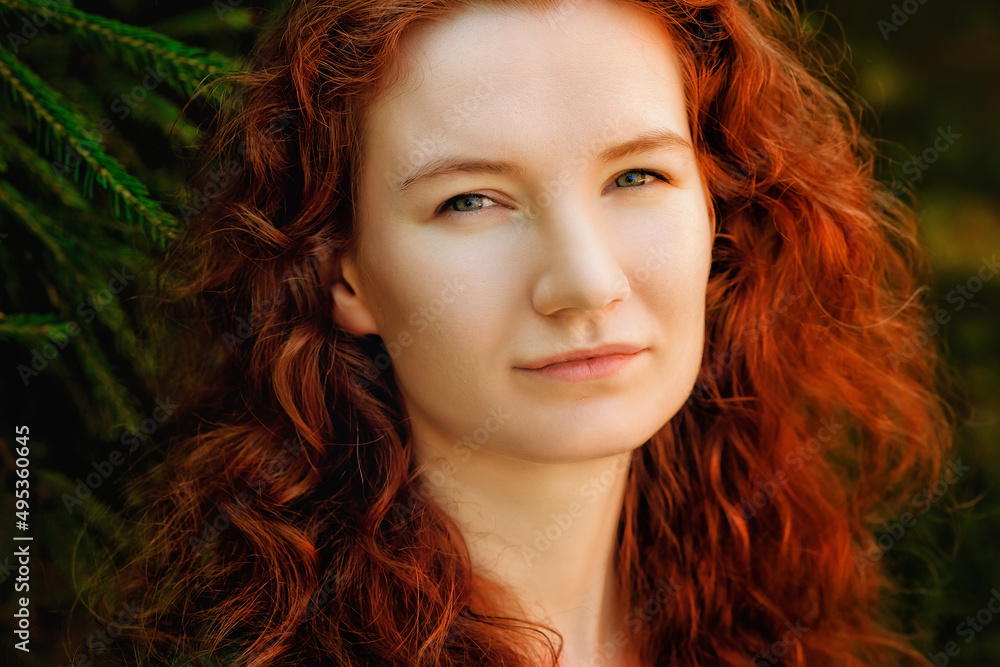 Portrait of a red-haired woman in the park. A curly-haired woman near the green fir branches. Portrait of a red-haired woman in the park. A curly-haired woman near the green fir branches.