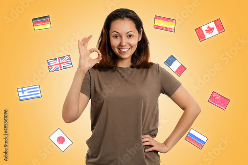 English language Day. A young smiling woman makes an OK gesture. Yellow background with flags of different countries. The concept of learning foreign languages photo