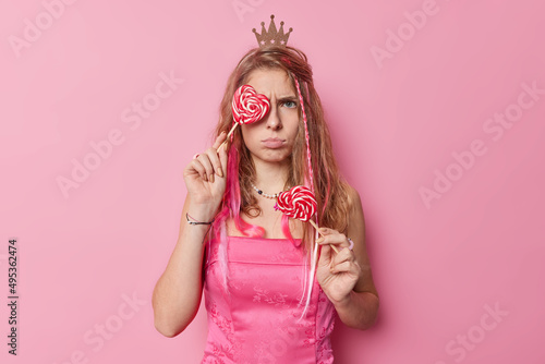 Horizontal shot of discontent young woman with long hair wears small crown and dress holds heart lollipops has sulking expression isolated over pink background. Angry princess with caramel candies