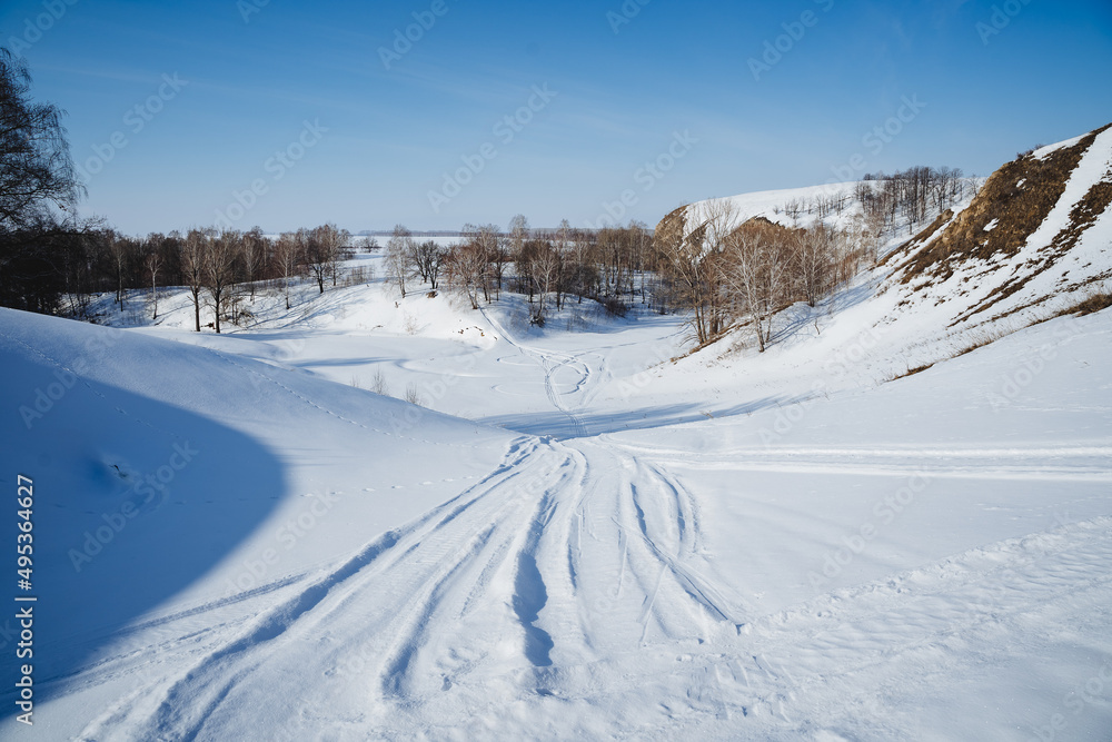 Snowmobile tracks on white snow, the road passes through a snow-covered river, winter landscape, sunny weather clear sky, winter cold