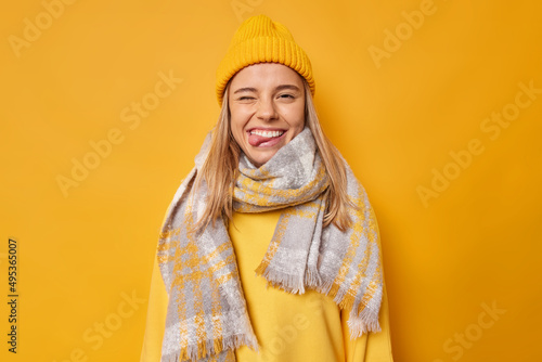 Positive carefree young woman winks eye and sticks out tongue wears hat casual jumper scarf around neck foolishes around isolated over yellow background goes crazy. Human facial expressions. photo
