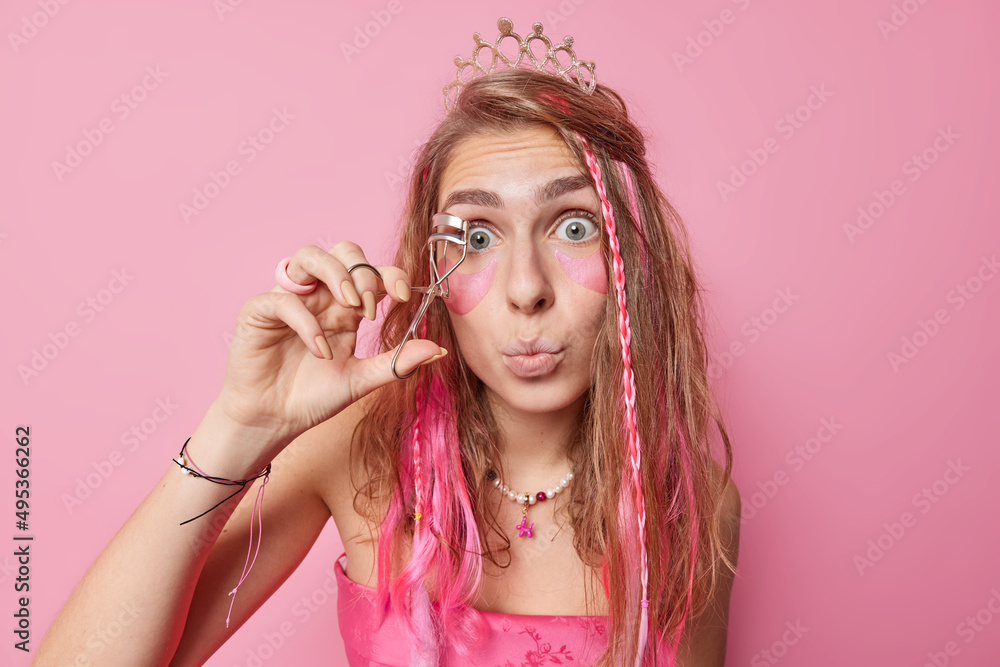 Surprised European woman prepares for party uses eyelashes curler applies makeup beauty hydrogel patches under eyes keeps lips rounded wears crown and festive dress isolated over pink background. Surprised European woman prepares for party uses eyelashes curler applies makeup beauty hydrogel patches under eyes keeps lips rounded wears crown and festive dress isolated over pink background.