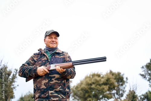 Male hunter in camouflage outerwear and cap carrying a gun while hanging out in the autumn field