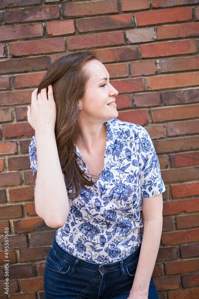 Portrait of brunette smiling woman standing in front of brick wall and stoking her hair, wearing printed blouse and jeans Portrait of brunette smiling woman standing in front of brick wall and stoking her hair, wearing printed blouse and jeans