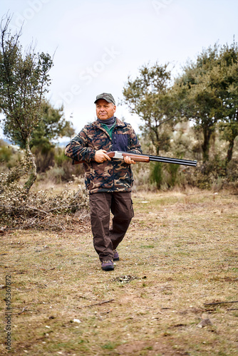 Full body aged male hunter in camouflage outerwear and cap carrying gun and looking away while spending time in autumn countryside