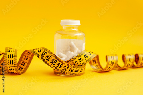 Tablets in capsules in a glass jar wrapped with a centimeter tape on a yellow background, horizontal shot. Weight loss concept, fat burner, vitamins, sports nutrition