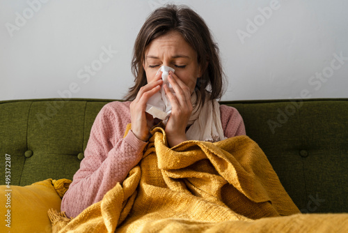 Sick woman blowing nose in facial tissue sitting on sofa at home photo