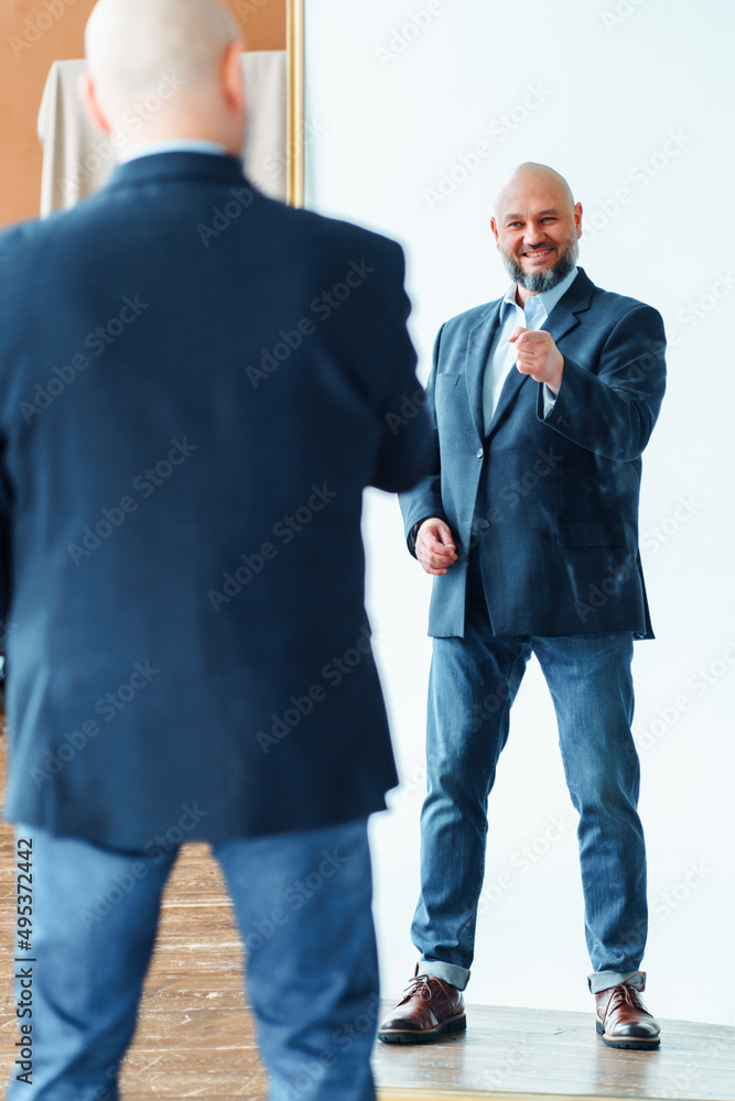 Portrait of happy middle-aged bald man with gray beard wearing blue suit jacket, pointing finger at mirror reflection.