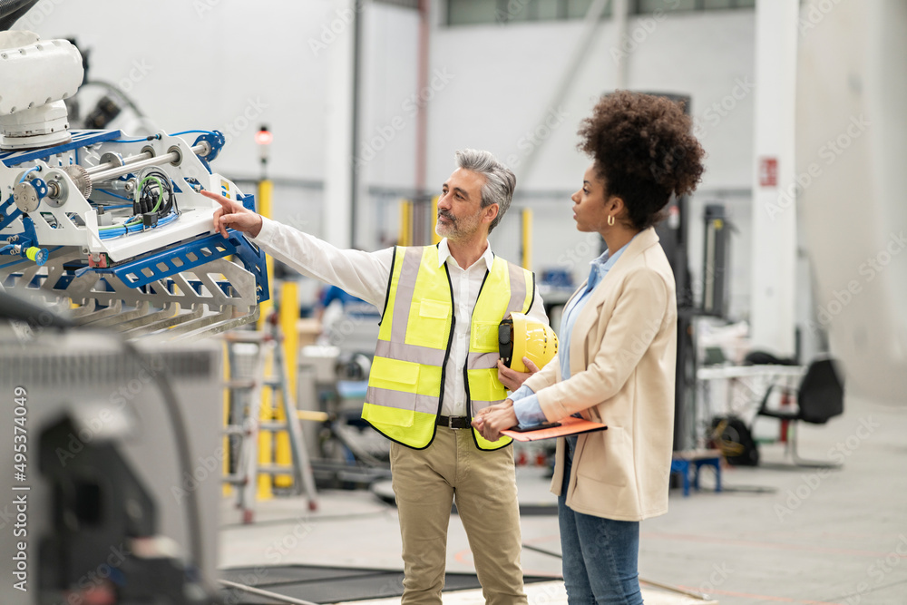 Engineer pointing at robotic arm discussing with businesswoman holding file folder in factory Engineer pointing at robotic arm discussing with businesswoman holding file folder in factory