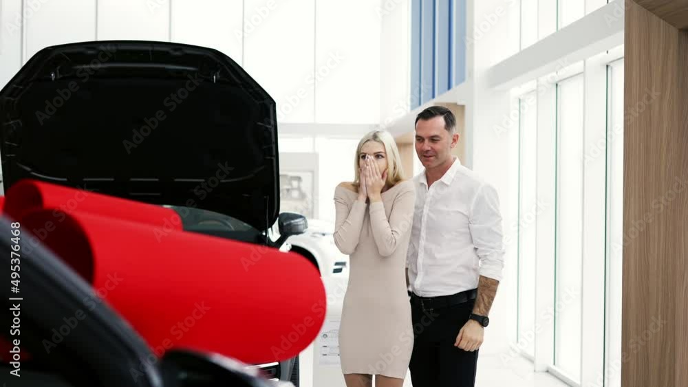 A man make surprise to a blonde woman, a new car with red bow in a showroom A man make surprise to a blonde woman, a new car with red bow in a showroom