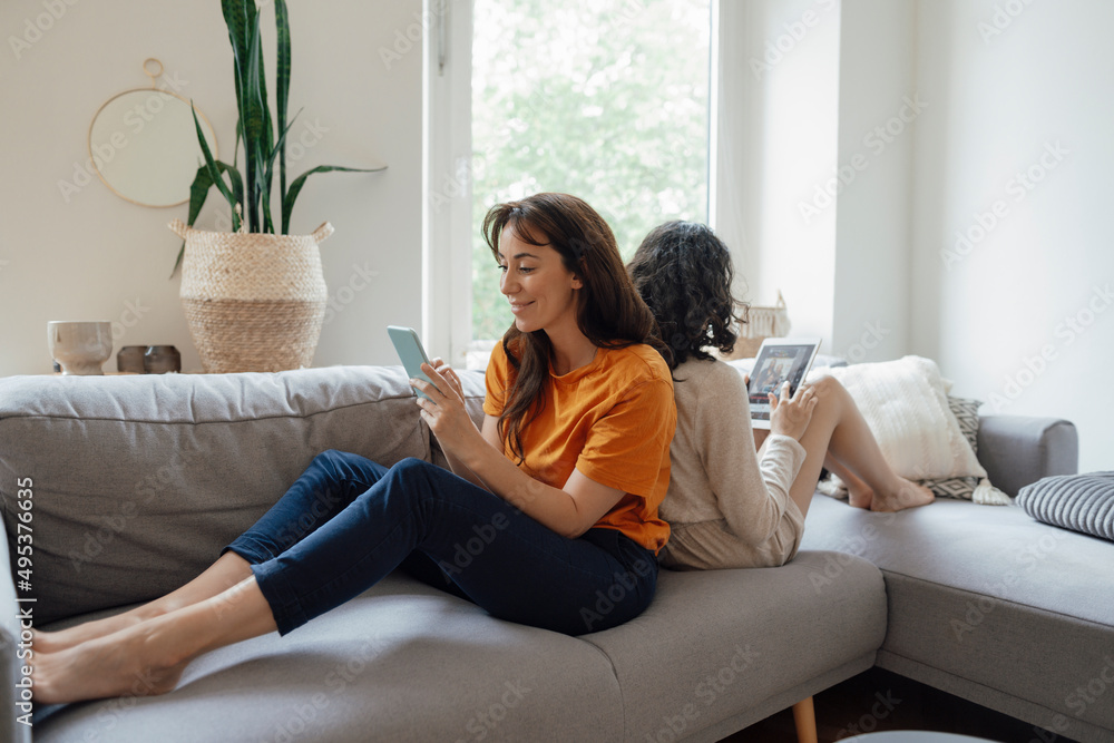Smiling woman using smart phone sitting back to back with daughter using tablet PC on sofa at home Smiling woman using smart phone sitting back to back with daughter using tablet PC on sofa at home
