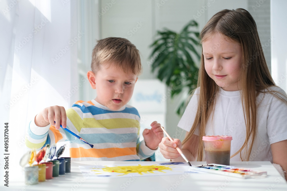 Fototapeta premium Brother and sister draw a picture with paints on a piece of paper. Children are learning to draw. Home creativity.
