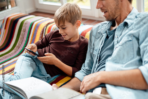 Man with book looking at son using smart phone sitting on sofa ta home photo