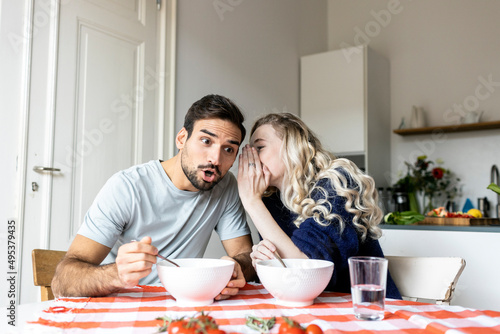 Surprise man listening to girlfriend whispering in ear sitting on dining table at home photo