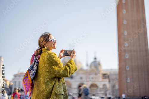 Young woman taking photo of famous bell tower of saint Mark basilica in Venice. Concept of visiting italian landmarks and travel. Woman wearing coat and shawl in italian style
