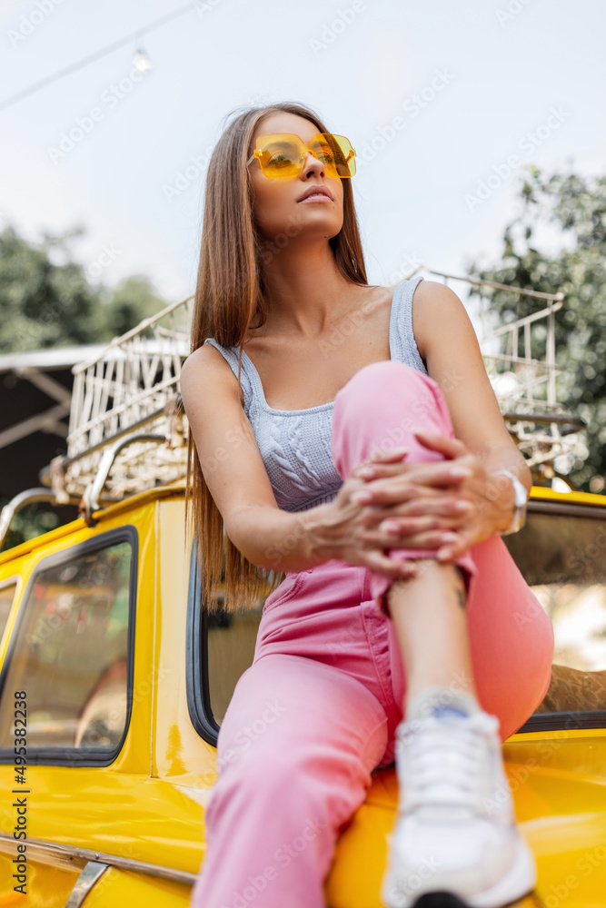 Cool fashion glamour young woman with yellow sunglasses in trendy tank top and pink jeans with shoes sits on yellow colored vintage car