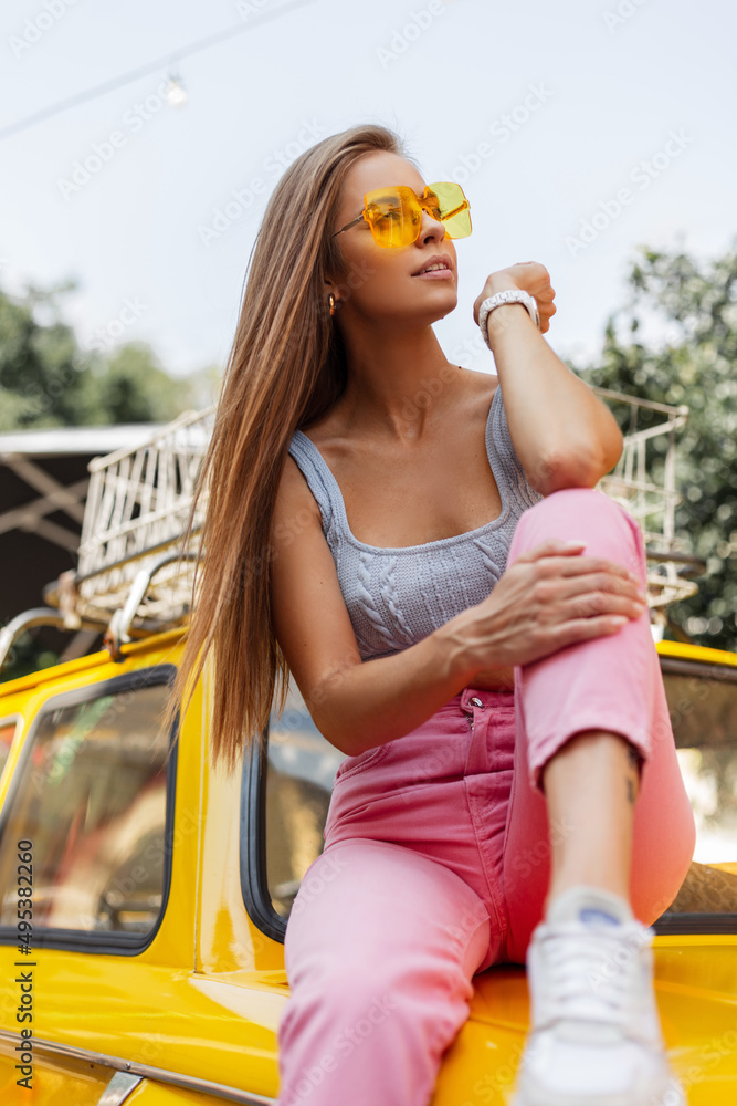 Beautiful young hipster woman in fashionable summer clothes with a sexy top, pink sneakers and yellow sits and rest on a car. Female summer colorful look outfit Stock-foto