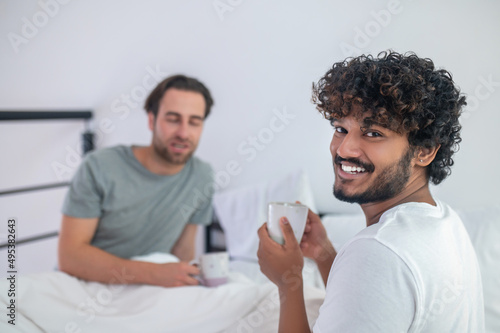 Man drinking coffee with his sleepy boyfriend in the bedroom