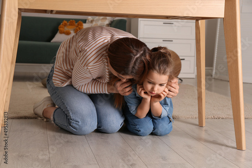 Scared mother with her little daughter hiding under table in living room during earthquake