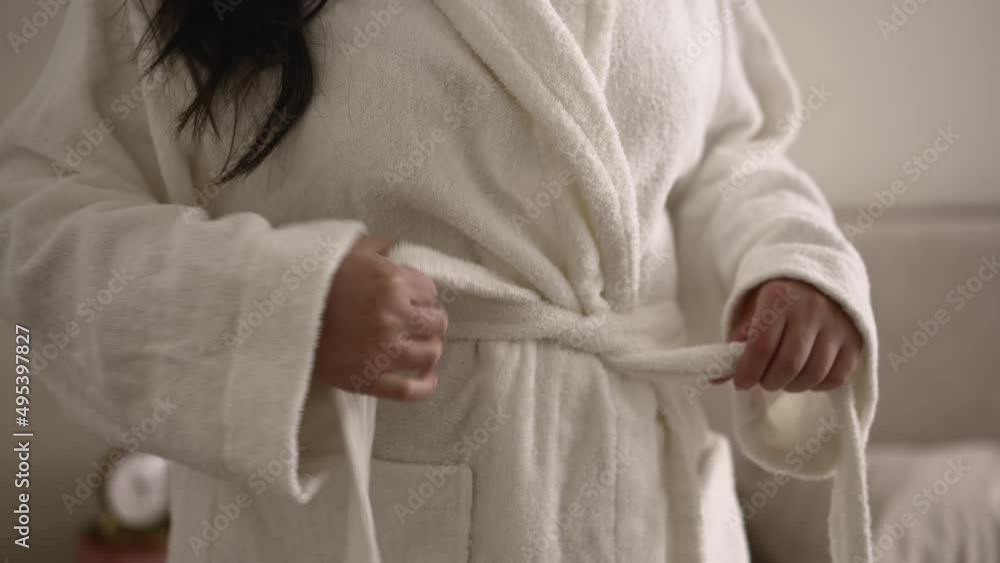 Closeup view of young overweight woman putting on bathrobe and standing in home room spbd. 4k Caucasian female puts on soft white robe over her body and poses after shower, stands in light interior