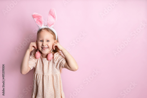 Happy Easter. A cute beautiful girl with bunny ears holds green eggs in her hands as earrings. Pink background. Space for text