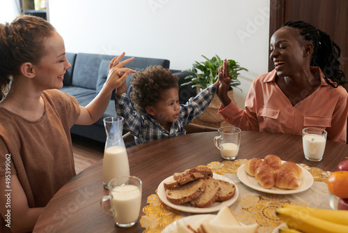 Happy diverse mothers giving high five to their little son when sitting at kitchen table with cup of milk
