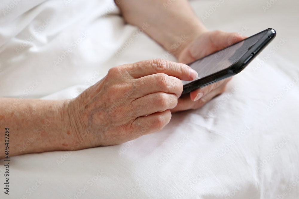 Elderly woman with smartphone in a bed, mobile phone in wrinkled female hands close up. Concept of online communication in retirement, sms, social media