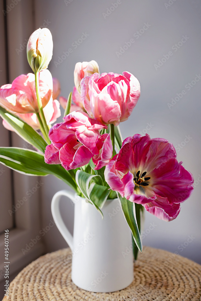 Obraz premium Pink open tulip flower blooming bouquet with green fresh stems in a white porcelain jar on a white wall background. Vibrant colourful botany floral home decor idea.