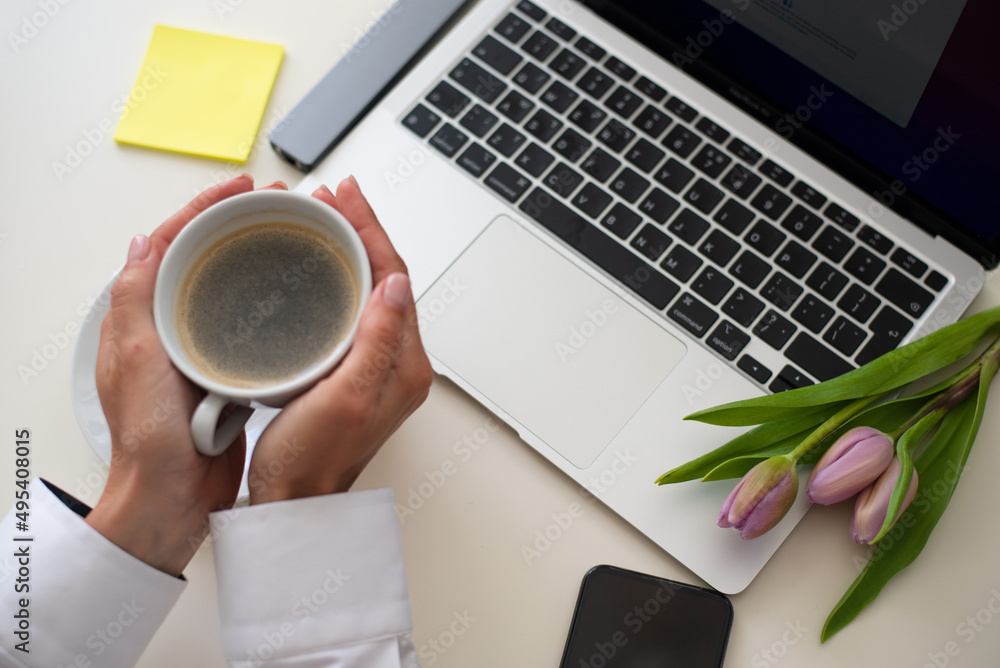 Woman's hands hold cup of coffee. Home office desk with laptop, smartphone, pink tulips on a white background. Flat lay Business workplace. Top view. businesswoman work on laptop Woman's hands hold cup of coffee. Home office desk with laptop, smartphone, pink tulips on a white background. Flat lay Business workplace. Top view. businesswoman work on laptop