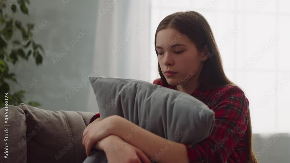 Sad young woman sits hugging pillow on couch in living room zoom in slow motion. Loss of beloved person overcoming. Psychotherapy help needing Sad young woman sits hugging pillow on couch in living room zoom in slow motion. Loss of beloved person overcoming. Psychotherapy help needing