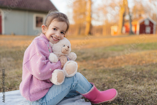 little girl with hearing aid sits on grass in knitted sweater and pink rubber boots with teddy bear, happy girl hug teddy bear otdoor photo
