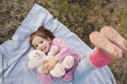 happy little girl in knitted sweater and rubber boots lies on grass with teddy bear and lift legs up