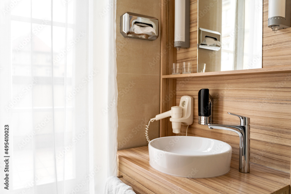 Fototapeta premium Stylish bright clean bathroom in hotel room with large window or door, sink, modern faucet, wooden furniture, hair dryer and mirror on sunny morning or day. Travel, holiday, vacation, interior design