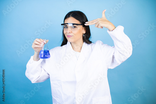 Young brunette woman wearing scientist uniform holding test tube over isolated blue background Shooting and killing oneself pointing hand and fingers to head like gun, suicide gesture.
