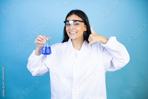 Young brunette woman wearing scientist uniform holding test tube over isolated blue background shouting with crazy expression doing rock symbol with hands up