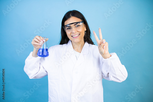Young brunette woman wearing scientist uniform holding test tube over isolated blue background showing and pointing up with fingers number two while smiling confident and happy