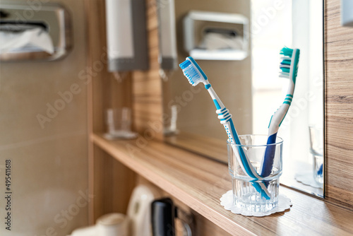 Two used blue and turquoise toothbrushes in glass cup stand on wooden shelf near mirror and window in bright modern clean hotel bathroom on sunny morning or day close up. Daily routine, healthcare