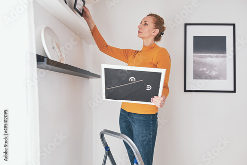 Obraz na plátně Woman on the ladder hanging pictures and photos on the shelf and wall at home