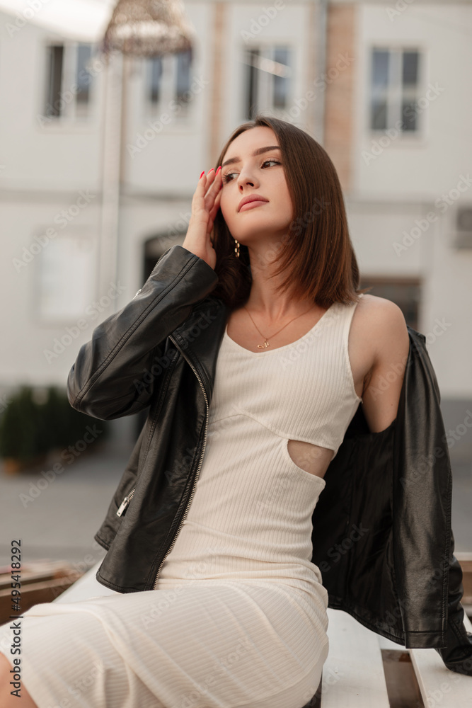 Cute beautiful Caucasian woman model in a fashionable black leather jacket and stylish white dress sits and relaxes in the city