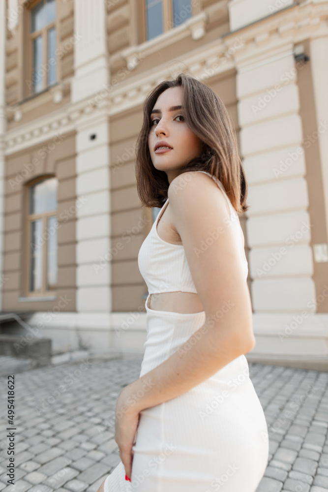 Model beautiful woman with a slender body and short hair in a fashionable white dress walking in the city and looking at the camera