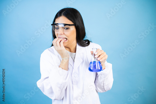 Young brunette woman wearing scientist uniform holding test tube over isolated blue background looking stressed and nervous with hands on mouth biting nails. anxiety problem.