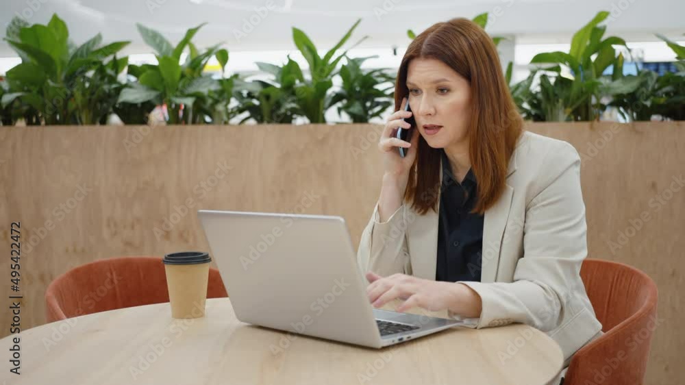Adult businesswoman talking on phone and typing on laptop in cafe, coffee cup beside her, green plants on background, entrepreneur working online, online business Adult businesswoman talking on phone and typing on laptop in cafe, coffee cup beside her, green plants on background, entrepreneur working online, online business
