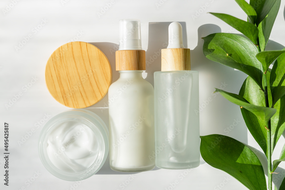 White cosmetic bottles and open jar with face cream on white background with green ruscus leaves. Skin care, body treatment, beauty concept. Unbranded packaging for skincare treatment