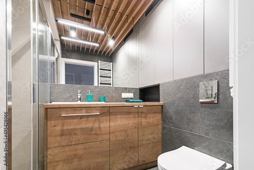 Łazienka z umywalką, baterią, dużym lustrem oraz kabiną prysznicową. Toaleta w mieszkaniu. Kolory szarości i drewno photo