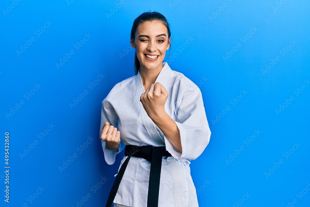 Beautiful brunette young woman wearing karate fighter uniform with black belt doing attack pose winking looking at the camera with sexy expression, cheerful and happy face. Beautiful brunette young woman wearing karate fighter uniform with black belt doing attack pose winking looking at the camera with sexy expression, cheerful and happy face.